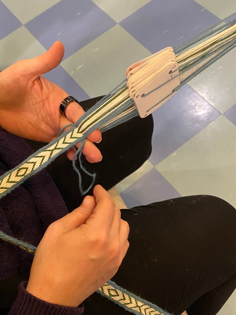 Kuvassa on lautanauhaloimi, kutojan kädet, aloitettu nauha ja valkoiset nauhalaudat loimeen pujotettuina.