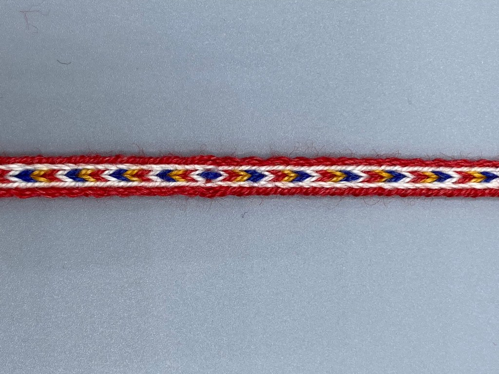 Punareunainen valkopohjainen lautanauha, jonka keskellä juoksee valko-puna-kelta-sininen nuolirivi.